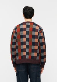 Άνδρας που φοράει πολύχρωμο πουλόβερ με καρό μοτίβο σε κόκκινο, ναυτικό μπλε, καφέ και γκρι τετράγωνα, συνδυασμένο με σκούρο μπλε τζιν παντελόνι, από πίσω.