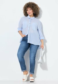 Camisa de botones a rayas azul claro con mangas largas, combinada con vaqueros azul oscuro y sandalias de cuña blancas de punta abierta, sosteniendo un pequeño bolso plateado.