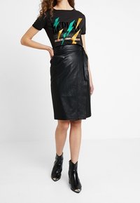Jupe enveloppante en cuir noir associée à un t-shirt noir avec des graphismes de foudre en vert, jaune et orange. Le mannequin porte des bottines noires.