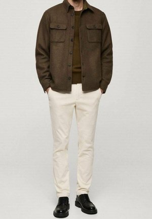 Mann trägt braune Hemdjacke mit Knöpfen, olivgrünen Pullover, cremefarbene Hose und schwarze Lederschuhe und steht mit den Händen in den Taschen.