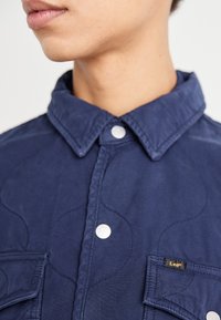 Chemise bleu marine avec un motif texturé, dotée d'un col, de deux poches avant et de fermetures à boutons argentés. Étiquette de la marque Lee sur la poche.