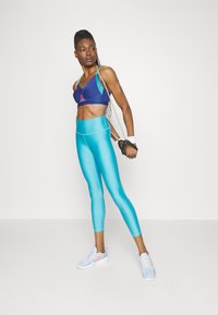 Sostén deportivo en azul y morado, combinado con leggings aqua. Ambos son ajustados y están hechos de un material suave y elástico. Zapatillas deportivas blancas con detalles en naranja.