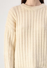 Knit cream sweater featuring a chunky ribbed texture, round neckline, and elongated sleeves with diagonal rib details.