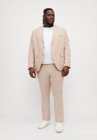 Traje de lino beige claro con chaqueta de un solo botón y pantalones a juego, combinado con una camiseta blanca y zapatillas blancas.