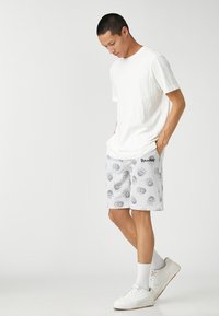 Camiseta de algodón blanca combinada con pantalones cortos grises que presentan patrones florales negros. El modelo lleva zapatillas blancas y calcetines hasta los tobillos.