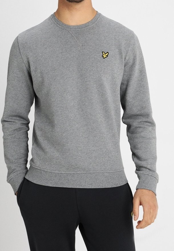 Homme portant un sweat-shirt gris à col rond avec un petit logo d'oiseau jaune sur la poitrine et un pantalon noir, mains dans les poches.