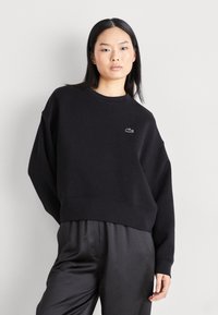 Musta huppari, jossa on pyöreä kaula-auko ja laskeutuvat olkapäät. Vasemmassa rinnassa pieni valkoinen Lacoste-logo. Pehmeä kangas.