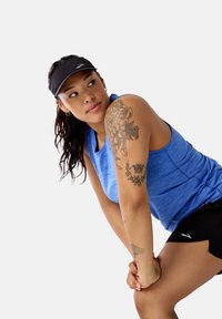 Sporta topiņš zilā krāsā, izgatavots no teksturēta auduma, kopā ar melnām šortiem. Ievērojams melns vizieris un redzami ziedu tetovējumi uz roku.