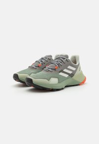 Trail running shoes in grey and mint green, featuring a textured upper, three white stripes, and orange accents on the laces and sole.