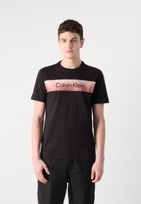 Svart bomull t-skjorte med en horisontal rosa gradientstripe og "Calvin Klein" tekstlogo i midten. Korting ermer og rund hals.