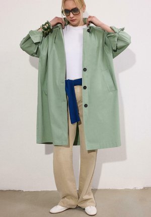 Nő lazán viselt világoszöld kabátot igazít, fehér inget, bézs nadrágot, derekán megkötött kék pulóvert, fehér cipőt, napszemüveget és zöld karkötőket visel.