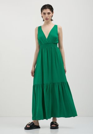 Woman wearing a sleeveless green maxi dress with a deep V-neck and black slide sandals with gold buckles, standing against a plain white background.