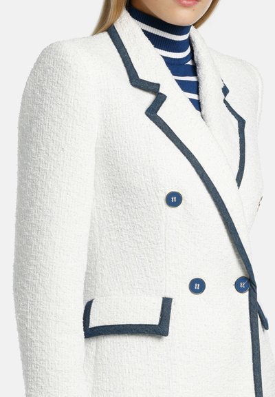 Witte textuurblazer met donkerblauwe accenten, dubbele borstsluiting, twee blauwe knopen en een zak met blauwe details.