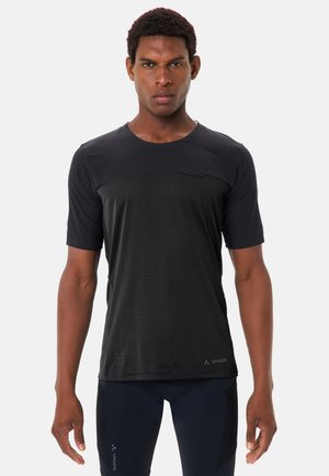 Junger Mann, der ein schwarzes, kurzärmeliges Sportshirt und passende Radsportshorts trägt, steht vor einem einfarbigen hellen Hintergrund.