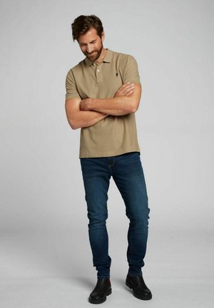 Beige polo paita lyhyillä hihoilla ja pienellä logolla, yhdistetty tummansinisiin farkkuihin ja mustiin saappaisiin. Istuvuus on rento ja muototettu.