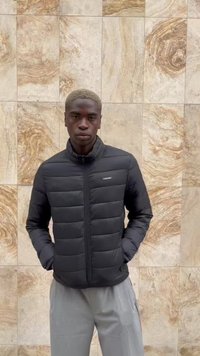 Un tânăr cu părul scurt și blond, îmbrăcat într-o geacă neagră din puf și pantaloni gri deschis, stă în fața unui perete din piatră crem, cu dale.