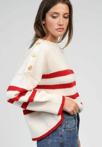 Pull en tricot blanc avec des rayures rouges, manches côtelées et ourlet, doté de boutons dorés sur l'épaule. Associé à un jean en denim bleu.
