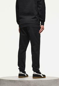 Persoon in zwarte joggingsbroek, zwart sweatshirt en zwart-witte sneakers staat op een rond platform tegen een effen achtergrond.