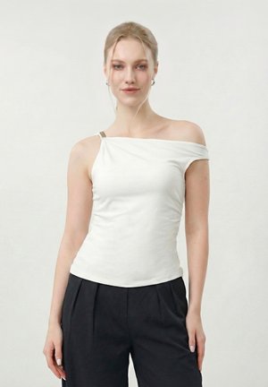 Young woman wearing a white asymmetrical off-shoulder top and black pants, standing against a plain light background.