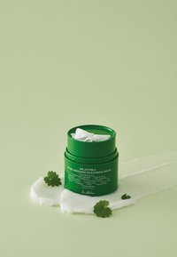 Zielony okrągły pojemnik z balsamem oczyszczającym z białą wewnętrzną pokrywką, otoczony kremową konsystencją i małymi zielonymi liśćmi na miękkim zielonym tle.