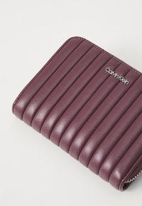 Ljubičasti kožni novčanik s okomitim rebrastim uzorkom, koji na prednjoj strani ima srebrni logo Calvin Klein i zatvaranje na zip.