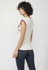 T-shirt blanc avec motif floral, accents rouges aux épaules, coupe ajustée, manches courtes ; associé à un jean bleu foncé. Texture de tissu lisse.
