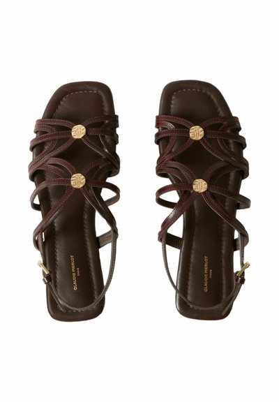 Coppia di sandali bassi in pelle marrone scuro con cinturini incrociati e dettagli circolari dorati, punta quadrata e fibbie alla caviglia.