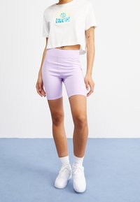 Lyhytvalkea t-paita, jossa on sininen teksti, istuvien laventelinväristen shortsien päällä. Mallilla on päällään valkoiset lenkkarit, joissa on nilkkasukat, ja hän seisoo sinisellä pinnalla.