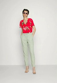 Mulher vestindo uma blusa curta de mangas com padrão floral vermelho, calças de cordão verde claro, sapatos de salto brancos e óculos de sol cor-de-rosa, de pé com as mãos nos bolsos.