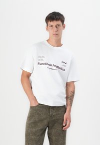White cotton t-shirt with a round neckline and short sleeves, featuring printed text and logos in dark purple. Worn with olive green textured pants.