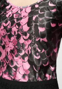 Κοντινό πλάνο εφαρμοστής μπλούζας με ροζ και μαύρο μοτίβο που μοιάζει με λέπια και λεπτομέρεια κειμένου "Versace Jeans Couture" στο ύφασμα.