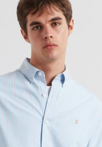 Lichtblauw overhemd met knopen, met witte en roestkleurige strepen, voorzien van een kraag en een klein geborduurd logo op de linkerborst.