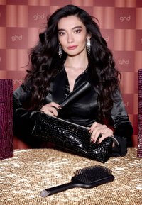 Czarna lakierowana kopertówka z teksturą krokodyla, trzymająca czarną prostownicę do włosów. Na złotej serwetce leży czarna szczotka do włosów z szerokim uchwytem.