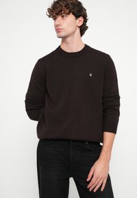 Csokoládébarna kötött pulóver kerek nyakkal és bordázott mandzsettákkal, kis logóval a mellrészen, fekete farmerrel párosítva.