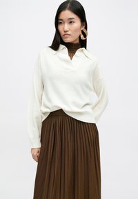 Κρέμα πουλόβερ με V ντεκολτέ και ριγωτό γιακά, συνδυασμένο με μια πλισέ καφέ φούστα. Μεγάλες χρυσές κρεμαστές σκουλαρίκια προσθέτουν μια σημαντική λεπτομέρεια.