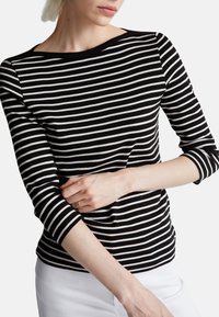 Czarna i biała bluzka w paski z rękawami 3/4 i szerokim dekoltem, wykonana z miękkiego materiału. Na dole widoczne białe spodnie.