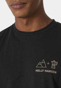 Černé bavlněné tričko s grafickým potiskem zlato barvy zobrazující hory a kávovar, s textem "HELLY HANSEN" níže.