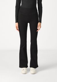 Black flared pants feature a stretchy, smooth fabric with a high waistband. Paired with white sneakers for a contrasting look.