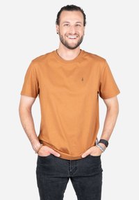Korte mouw, ronde hals t-shirt in roestrood katoen, met een klein boomlogo op de borst en een ontspannen pasvorm. G gedragen met donkere jeans.