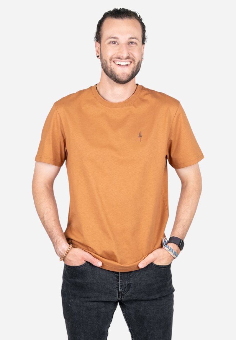 Korte mouw, ronde hals t-shirt in roestrood katoen, met een klein boomlogo op de borst en een ontspannen pasvorm. G gedragen met donkere jeans.