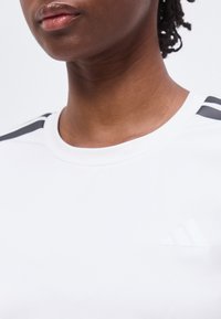 Gros plan d'une personne portant un t-shirt blanc Adidas avec des bandes noires aux épaules et un logo Adidas discret sur la poitrine.