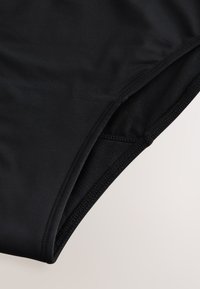 Gros plan sur un tissu noir montrant une partie d’un vêtement avec une couture courbe et un matériau lisse et extensible, probablement un bas de maillot de bain ou un vêtement de sport.