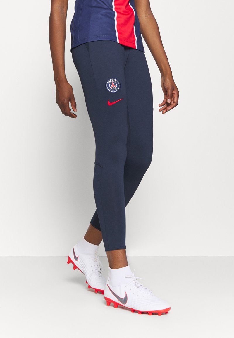 Personne portant des leggings bleu marine Paris Saint-Germain et des chaussures de football blanches avec des crampons rouges et des logos Nike.