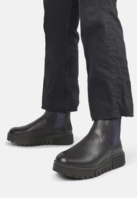 Czarne skórzane botki za kostkę z grubą gumową podeszwą, z elastycznymi panelami bocznymi i gładkim wykończeniem. Noszone z ciemnymi, zwężanymi spodniami.