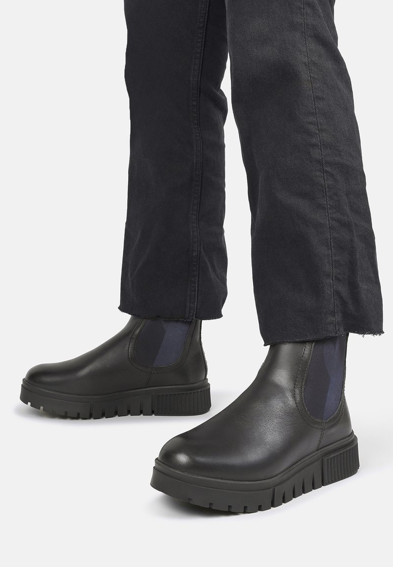 Czarne skórzane botki za kostkę z grubą gumową podeszwą, z elastycznymi panelami bocznymi i gładkim wykończeniem. Noszone z ciemnymi, zwężanymi spodniami.