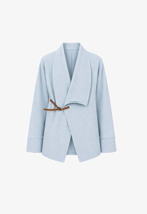 Femme portant une veste oversize bleu clair avec une boucle marron, une chemise blanche, un jean à jambes larges, des talons plateformes noirs et tenant un sac à main tissé.