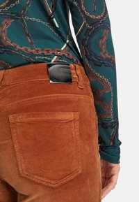 Braune Cordhosen mit aufgesetzten Taschen und einem glänzenden schwarzen Logo-Tag; getragen mit einem grünen Hemd mit Kettenmuster.