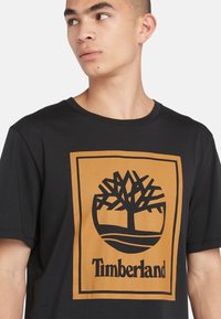 Zwarte katoenen T-shirt met een groot bruin en zwart Timberland boomlogo. Korte mouwen en een ronde halsontwerp. Gladde textuur.