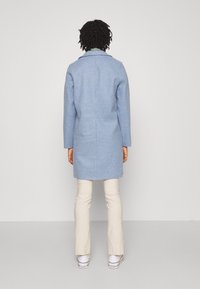 Cappotto in lana azzurro chiaro con taglio dritto, maniche lunghe e collo a punta, abbinato a pantaloni color crema e sneakers bianche.
