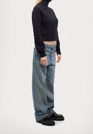 Femme portant un pull à col roulé noir texturé, un jean bleu clair ample et des chaussures noires épaisses, debout sur un fond uni.